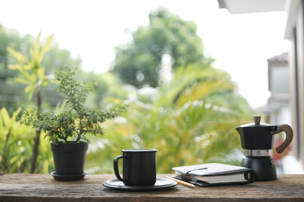 taza de café negra y olla de moka y olla de planta negra y diario de cuaderno en una vieja mesa de madera marrón