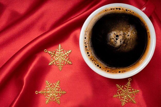 Una taza de café negro con adornos de estrellas de Navidad sobre un fondo de tela de seda de color rojo brillante.