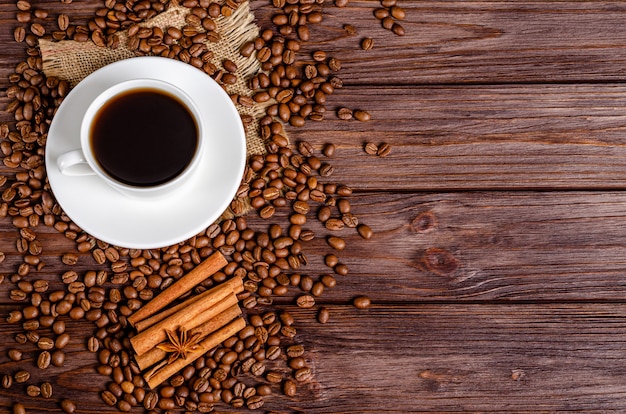 Una taza de café negro aromático en una taza blanca con granos de café tostado arábica, estrellas de anís, palitos de canela sobre un fondo de madera