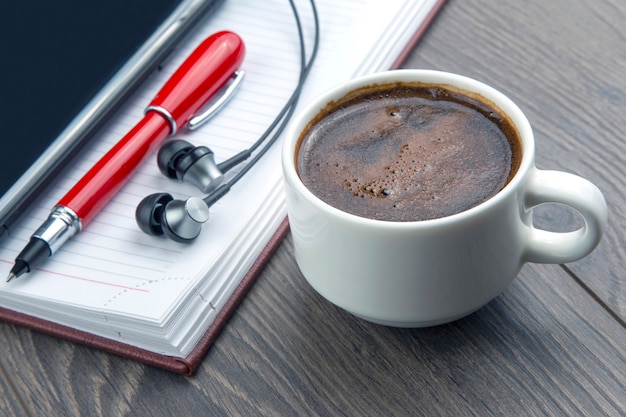 Taza de café negro, bolígrafo, teléfono móvil, portátil y auriculares sobre la mesa