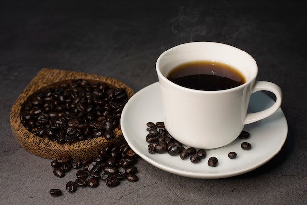 Una taza de café negro caliente sobre un fondo negro con granos de café tostados y vapor caliente que sale de la taza.