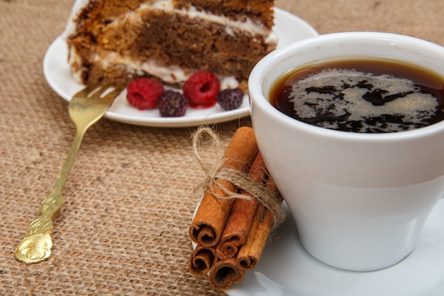 Taza de café negro, canela, tenedor, rebanada de bizcocho decorado con crema batida y frambuesas en la mesa con tela de saco.