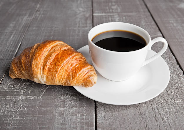 Taza de café negro y croissant para el desayuno en la superficie de madera