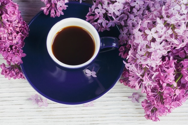 Taza de café negro, flores lilas sobre mesa de madera clara