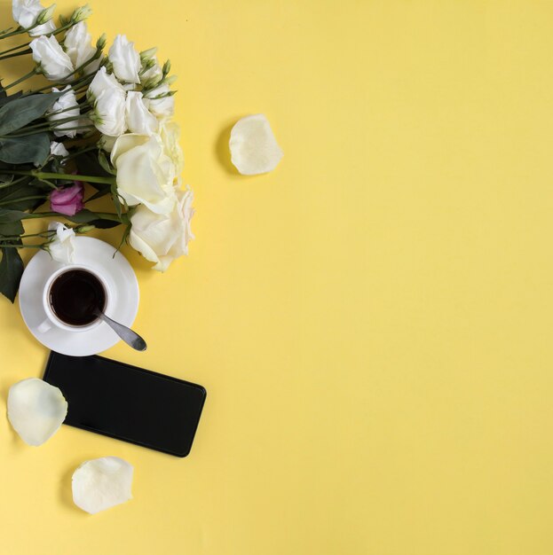 Taza de café negro con flores y smartphone sobre fondo amarillo con espacio de copia