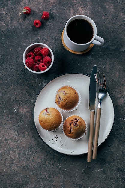 Taza de café negro y muffinf de frambuesa en placa sobre mesa de hormigón oscuro. Vista superior