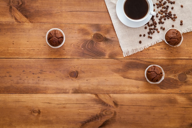 Taza de café negro, muffins y granos de café esparcidos sobre la mesa de madera marrón