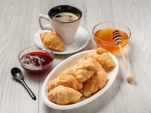 Taza de café negro con platillo, croissants recién hechos en plato de porcelana blanca, miel y mermelada de fresa en cuencos de vidrio con fondo de madera gris