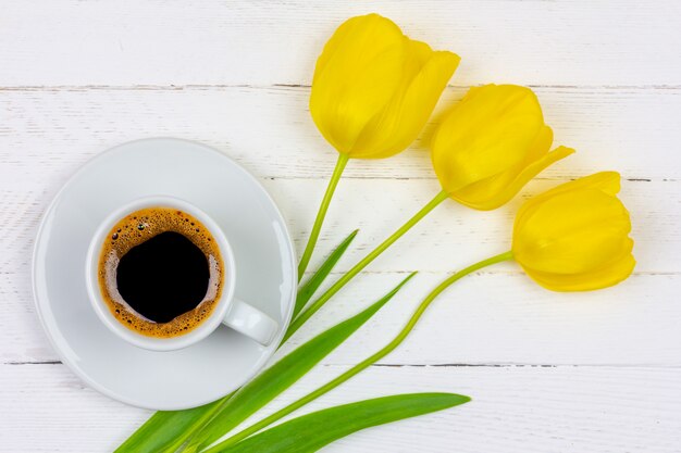 Una taza de café negro en un platillo y una flor de tulipán amarillo tres sobre un fondo de madera blanco