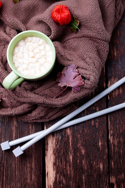 Taza de café o chocolate caliente con malvavisco cerca de una manta tejida y agujas.