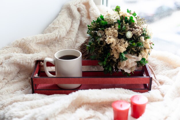 Una taza de café o té, flores secas y velas blancas y rojas, una caja de madera roja sobre una manta beige.