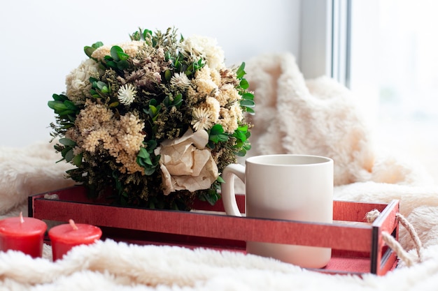 Una taza de café o té, flores secas y velas blancas y rojas, una caja de madera roja sobre una manta beige.