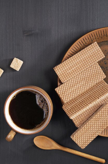 Taza de café y obleas de chocolate en un plato sobre fondo de madera negra, vista superior con espacio para copiar