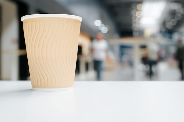 Taza de café de papel desechable se encuentra en la mesa blanca en el café, desenfoque de fondo claro lugar público, en el interior. Enfoque selectivo en taza con bebidas, espacio de copia. Vista lateral a nivel de objeto.