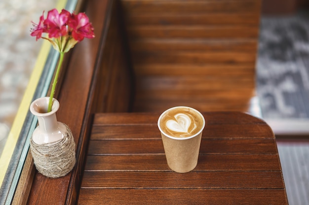 Una taza de café de papel con espuma en forma de corazón y una jarra con una flor roja sobre la mesa