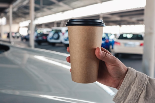 Una taza de café de papel en una mano en el fondo de un auto en un estacionamiento