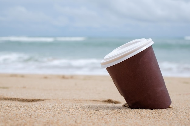 Taza de café de papel marrón en la playa de arena sobre el cielo nublado y el mar