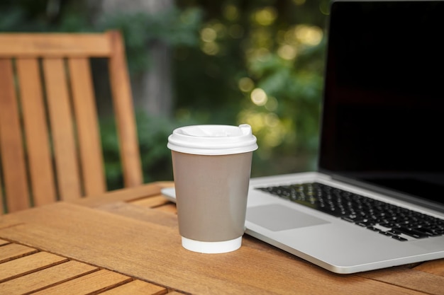 Una taza de café de papel y un ordenador portátil sobre una mesa de madera sobre un fondo de bosque borroso