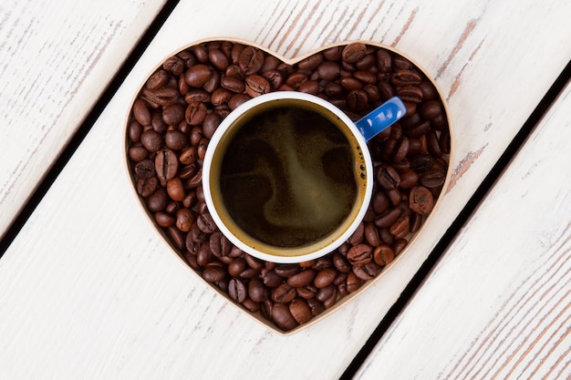 Taza de café de pie sobre el corazón de granos de café. Concepto de amor de café. Superficie de madera blanca.