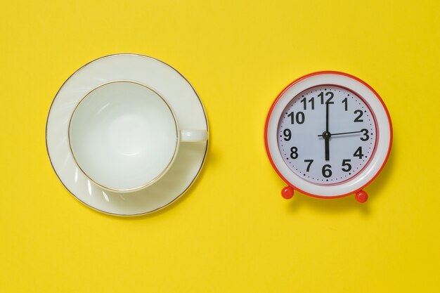 Una taza de café y un platillo y un despertador rojo sobre un fondo amarillo. El concepto de levantar el tono por la mañana. Endecha plana.