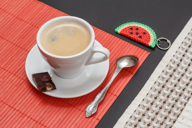 Taza de café en el platillo con un trozo de barra de chocolate una cuchara una servilleta de bambú y una baratija