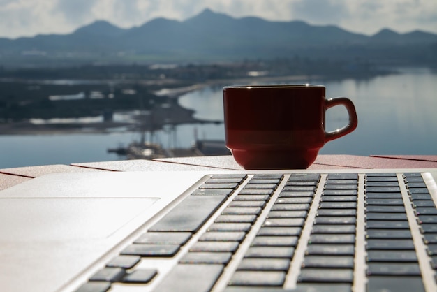 Taza con café en un primer plano de teclado tableLaptop rojo Vista del lago mar océano río