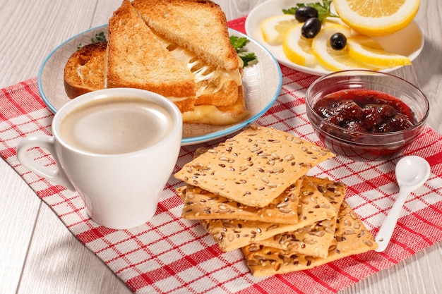 Taza de café, rebanadas de pan tostado con queso y perejil verde en plato blanco, tazón de vidrio con mermelada de fresa y cuchara con servilleta de cocina roja. Vista superior. buena comida para el desayuno.