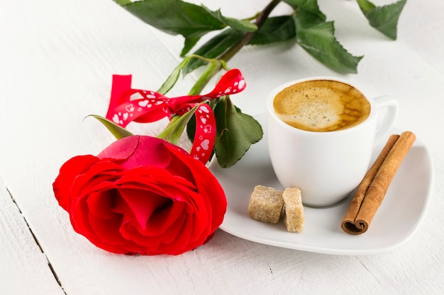 Taza de café, rosa roja, azúcar y canela sobre un fondo blanco de madera