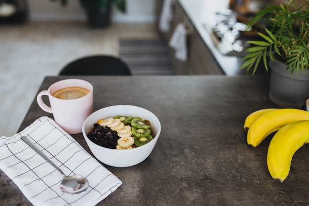 Taza de café rosa, tazón con frutas tropicales picadas, kiwi y plátano, arándanos, cuchara en barra de bar en la elegante cocina tipo loft