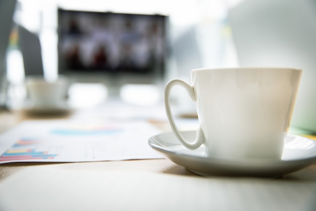 Taza de café sabrosa caliente blanca en un plato pequeño colocado en la mesa de trabajo con la computadora portátil en la sala de reuniones en el gráfico de la tasa de crecimiento objetivo frontal y el informe del gráfico en el monitor de pantalla grande en el fondo borroso.