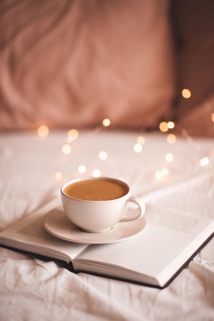 Taza de café sabroso en el libro de papel abierto en la cama sobre las luces de Navidad de cerca. Buenos días. Temporada de vacaciones de invierno. Hora del desayuno.