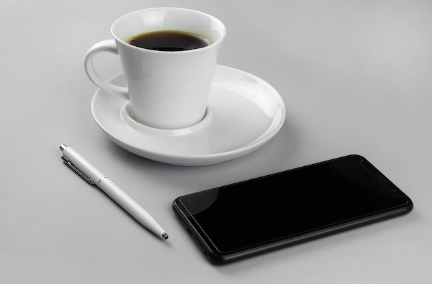 Taza de café, smartphone y bolígrafo blanco sobre un fondo gris.