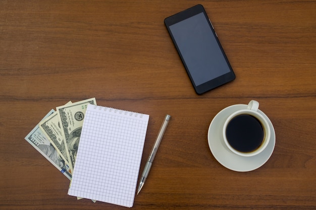 Taza de café, smartphone, dólares, bloc de notas y bolígrafo en el escritorio de madera