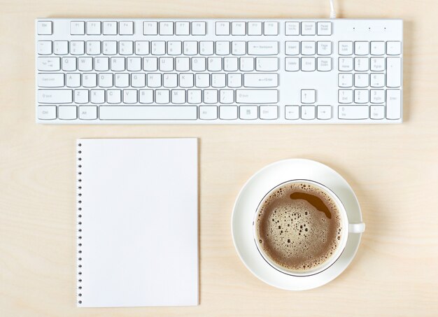 Taza de café, teclado y bloc de notas sobre superficie de madera clara.