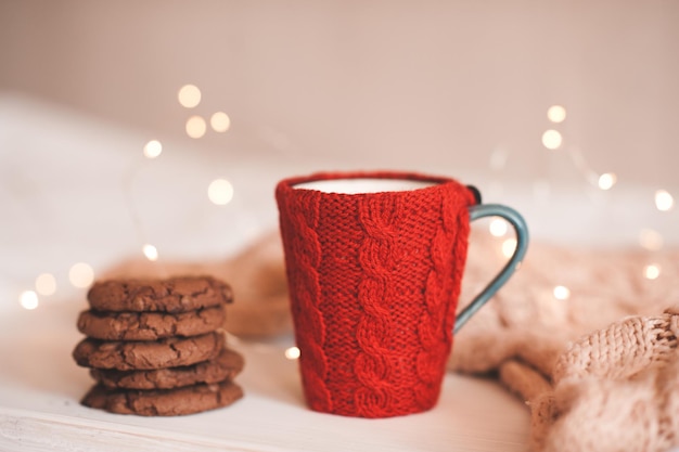 Una taza de café con tela de punto rojo con galletas de chocolate apiladas sobre las luces navideñas cerca de la cama