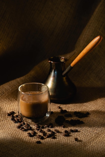 Una taza de café transparente con leche y un cezve sobre un fondo oscuro de arpillera