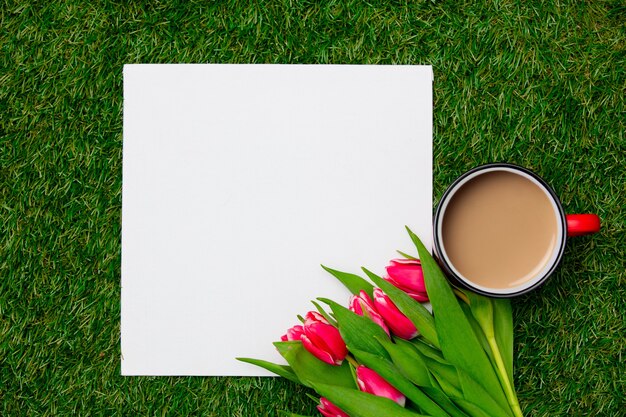 Taza de café y tulipanes cerca de cartón cuadrado sobre hierba verde