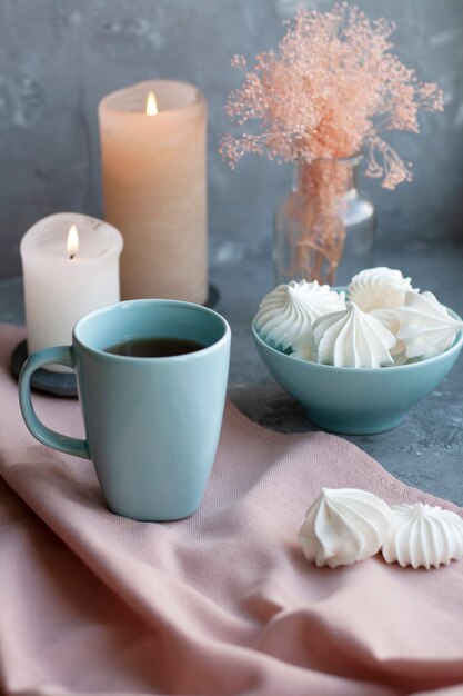 taza caliente de té negro con un tazón de merengues aireados sobre un mantel rosa, velas de cera, un jarrón de vidrio con hierbas decorativas