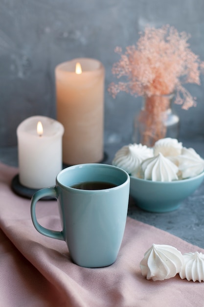 taza caliente de té negro con un tazón de merengues aireados sobre un mantel rosa, velas de cera, un jarrón de vidrio con hierbas decorativas