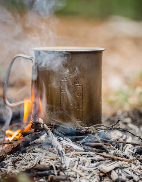 Taza de camping de metal con té caliente junto al fuego en el bosque