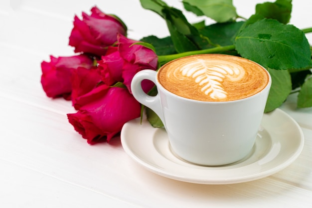 Taza de capuchino con arte y ramo de rosas en la mesa de madera de cerca