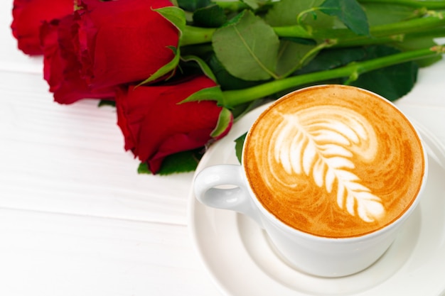 Taza de capuchino con arte y ramo de rosas en la mesa de madera de cerca