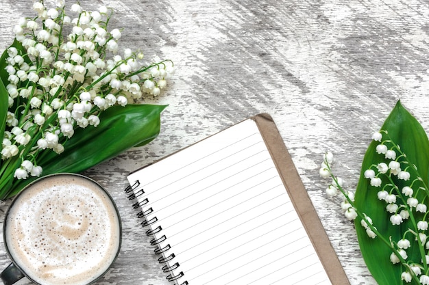 Taza de capuchino con flores de lirio de los valles de primavera y cuaderno forrado