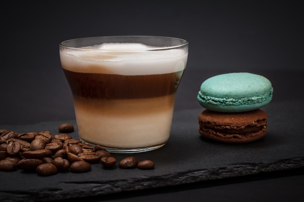 Taza de capuchino, granos de café y macarrones sobre tablero de piedra negra y fondo oscuro.