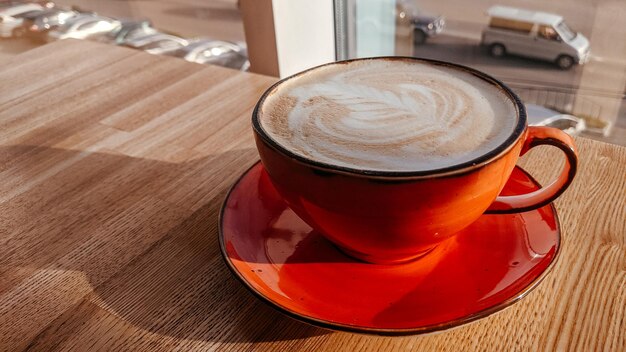 Una taza de capuchino rojo en una mesa de café con el telón de fondo de una ventana