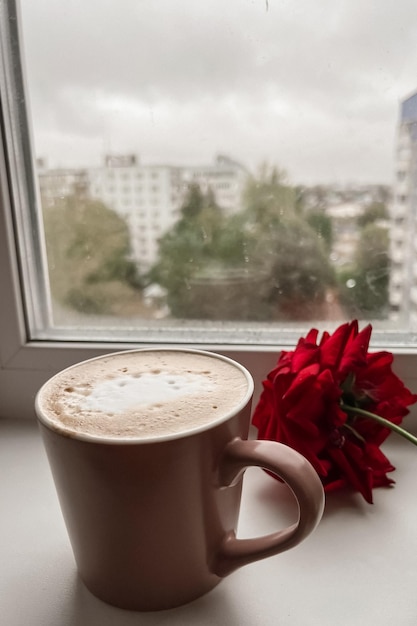 Una taza de capuchino rosa y una rosa de jardín roja están en el alféizar de la ventana Acogedor desayuno festivo en casa