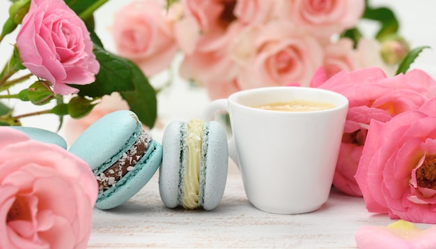 Taza de cerámica blanca con café y macarons sobre una mesa blanca, detrás de un ramo de rosas rosadas, cerrar