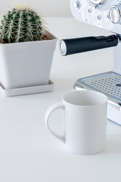 Taza de cerámica blanca de maqueta con máquina de café y cactus en mesa blanca en vista vertical.