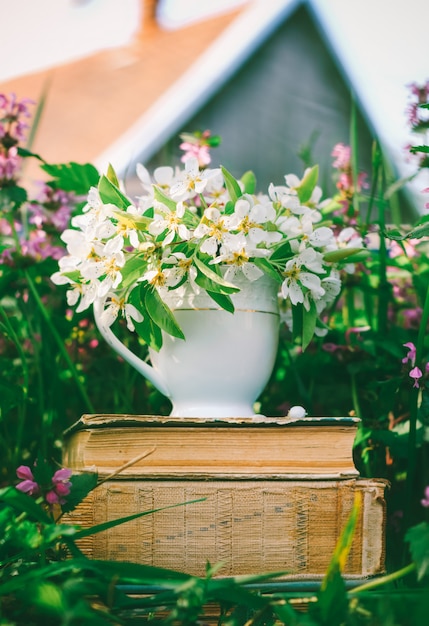 Taza de cerámica con flores blancas sobre una pila de libros antiguos en un claro con hierbas en flor en el contexto de una casa de pueblo