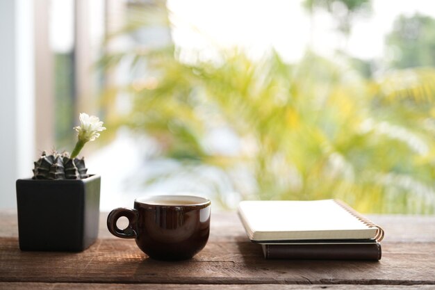 Taza de cerámica marrón café y flor de cactus blanco en una olla negra sobre una mesa de madera con cuaderno